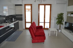 Relax in this stylish living space with a sofa, coffee table, and TV, surrounded by a potted plant and a kitchen area with a sink and wine bottle.