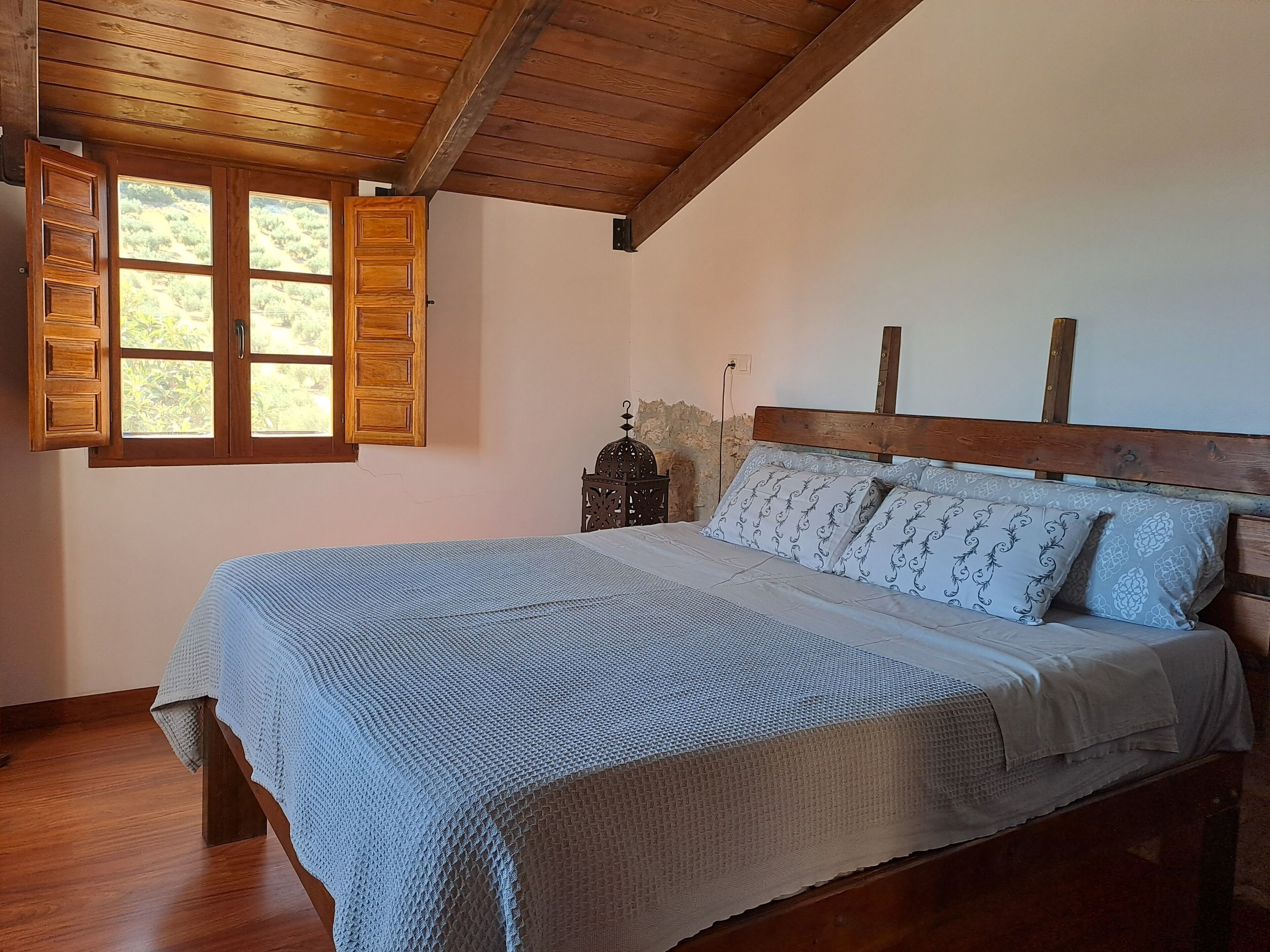 Une chambre avec un plafond en bois, un lit agrémenté d’oreillers à motifs, une table de chevet ornée d’un objet décoratif et une fenêtre avec des volets en bois.