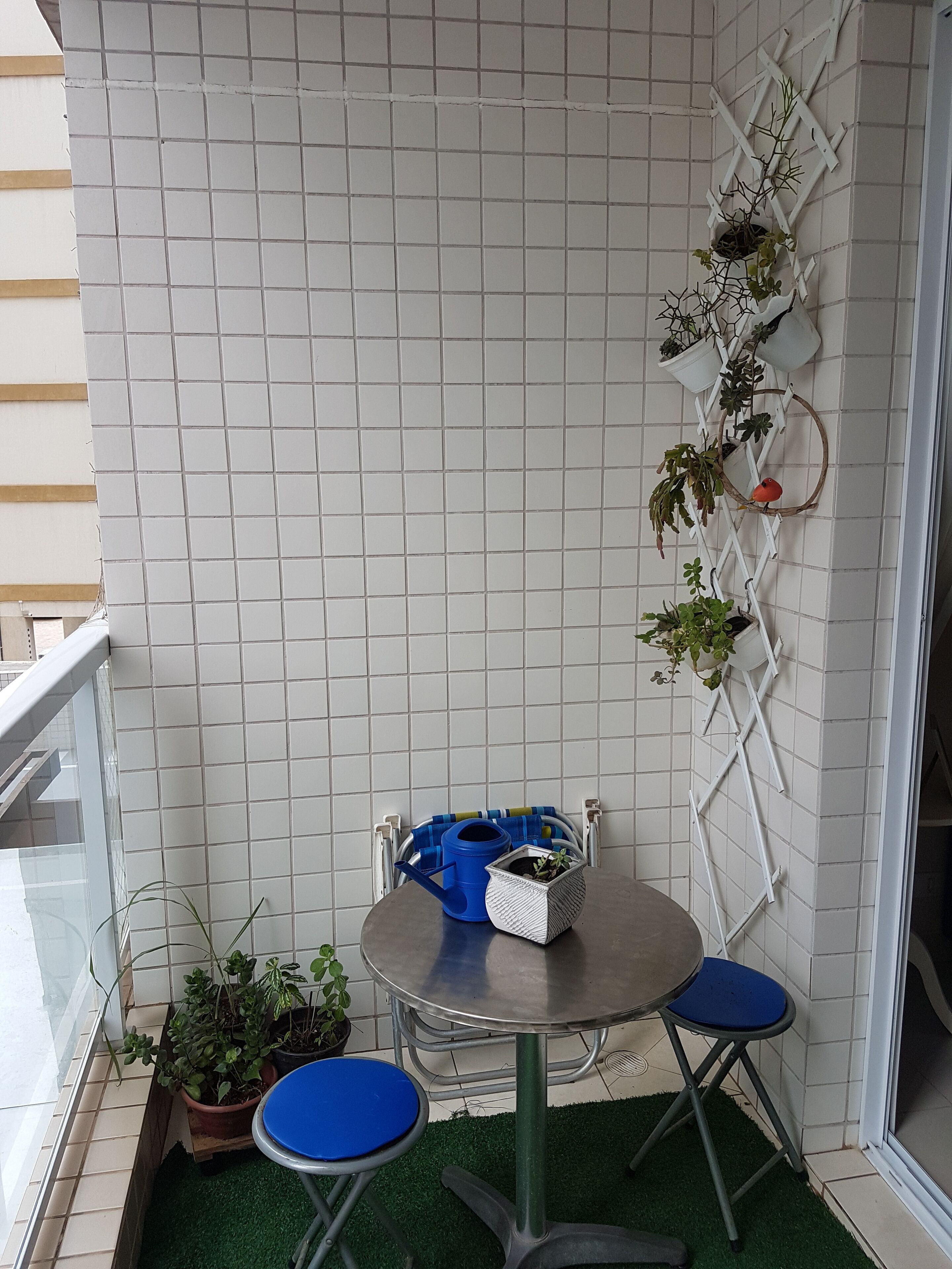 Un petit balcon avec un mur recouvert de carreaux, une chaise en métal, un tabouret bleu, une petite table avec un arrosoir bleu et un pot blanc, ainsi qu’un support vertical pour plantes, où sont disposées différentes plantes.