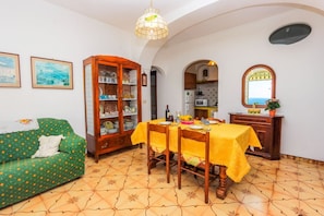 Un comedor con una mesa preparada para cuatro personas, un sofá verde, un mueble de madera con puertas de cristal, y una cocina visible a través de un arco.