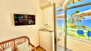 A room with a television mounted on the wall, a wicker chair with patterned cushions, a white storage unit, and a glass door leading to an outdoor area with a view of the sea and a tree. - Tramonto Praiano centre village (Praiano)