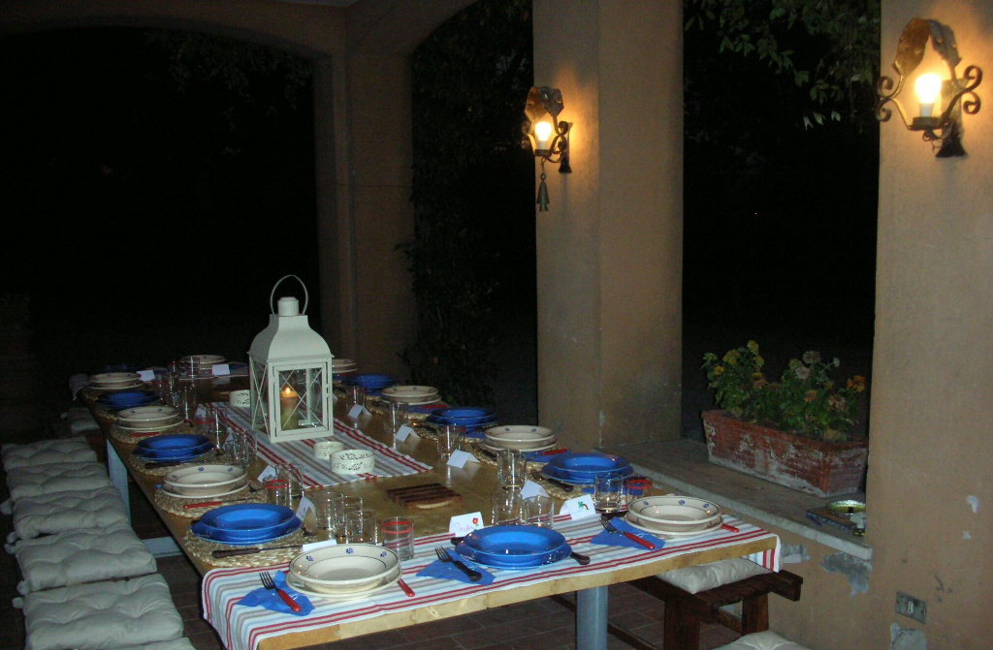 Une table dressée pour un repas en plein air, agrémentée d’une lanterne, de bougies et de lampes fixées au mur.
