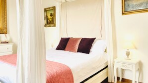 Een slaapkamer met een hemelbed, een nachtkastje met een lamp en een schilderij aan de muur.
