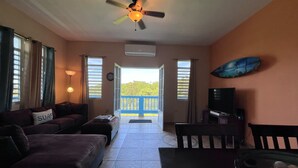 A living room with a sofa, a coffee table, a TV, and a ceiling fan. There is a balcony with a view of greenery.