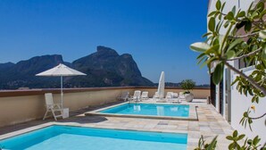 A rooftop pool with two separate sections, white lounge chairs, a white umbrella, and a view of mountains in the background. - Barramares Ocean View (Rio de Janeiro)