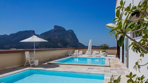 Una piscina en la azotea con dos zonas separadas, sillas de descanso blancas, una sombrilla blanca y vistas a montañas al fondo.