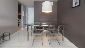 A modern dining area with a glass table and wireframe chairs, a pendant light, and a view into a laundry room. - Barramares Ocean View (Rio de Janeiro)
