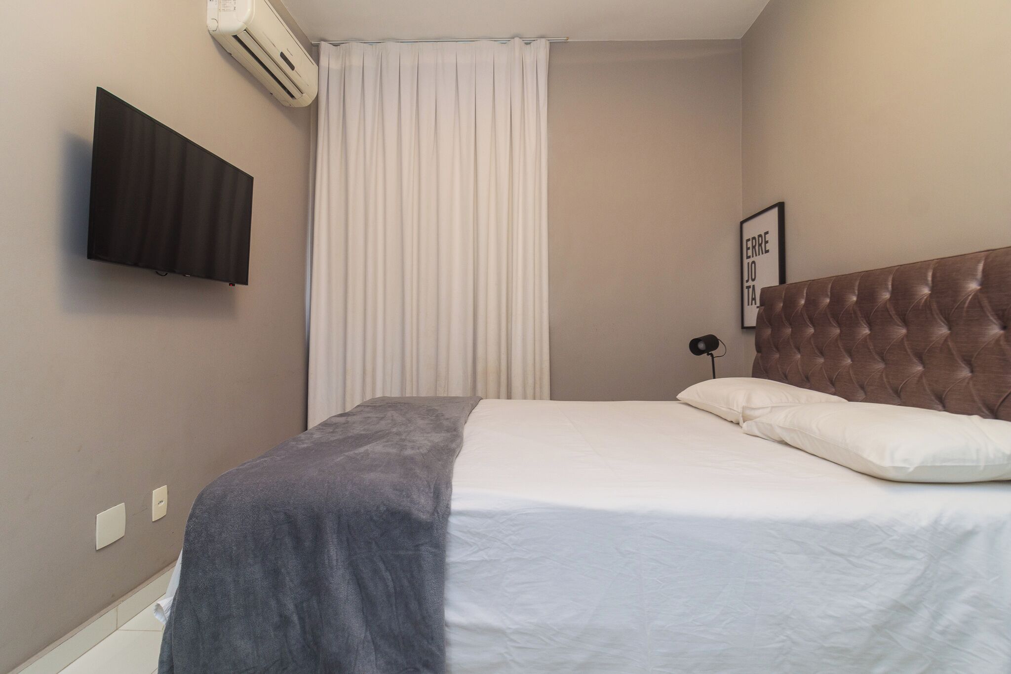 Une chambre à coucher avec un lit, un téléviseur fixé au mur, un climatiseur mural et un tableau encadré.