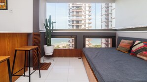 Un apartamento moderno con balcón, un cactus, una cama con almohadas coloridas y vistas a la playa y edificios altos.