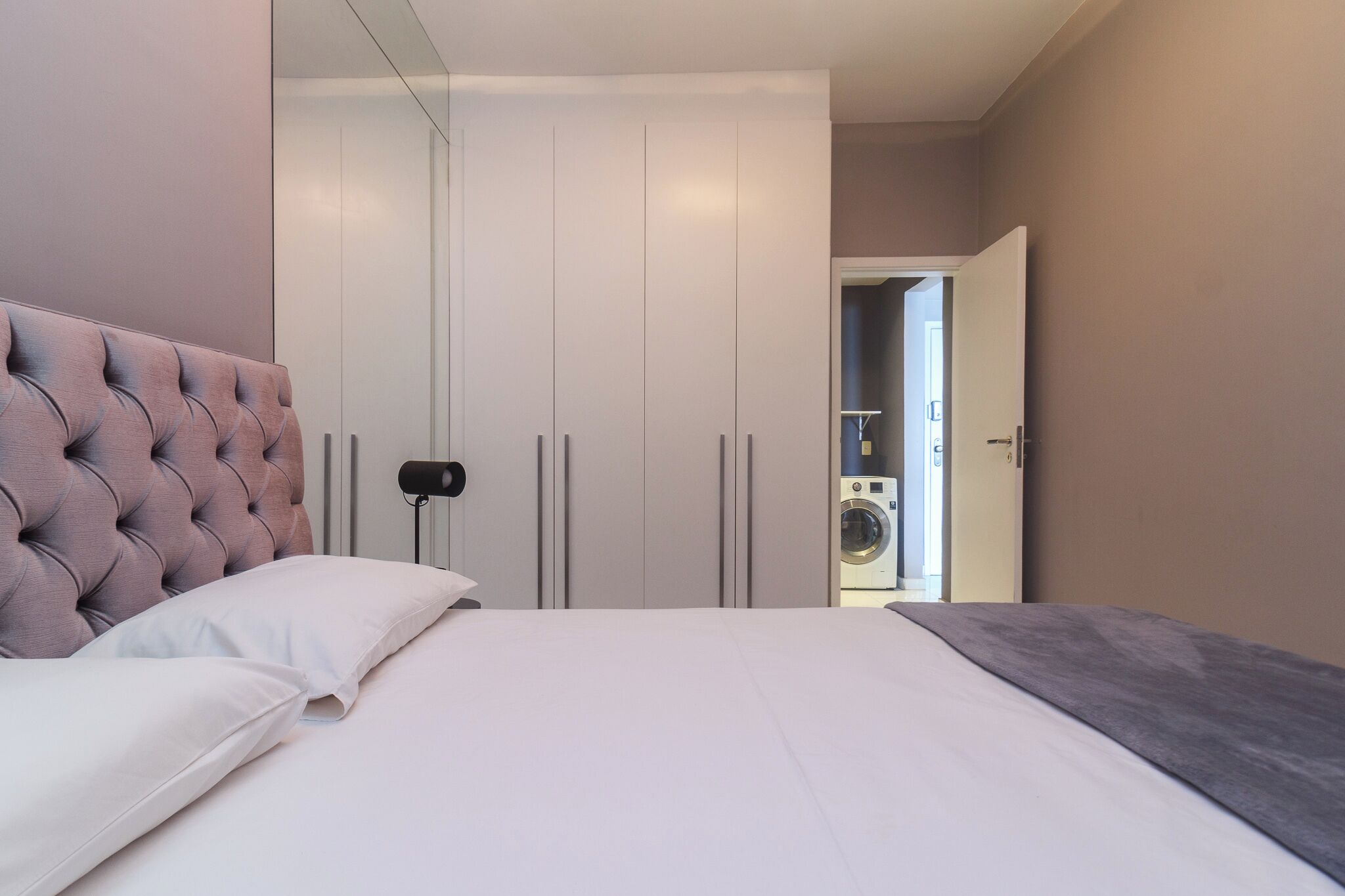 Un dormitorio con una cama grande, cabecera tapizada, armario con múltiples puertas y una lavadora al fondo.