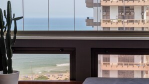 Una habitación con un amplio ventanal con vista a una playa, un cactus en maceta y una cama con cabecera de madera.