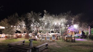 Un espace de restauration en plein air, avec des tables et des chaises disposées sous les arbres, illuminé par des guirlandes lumineuses.