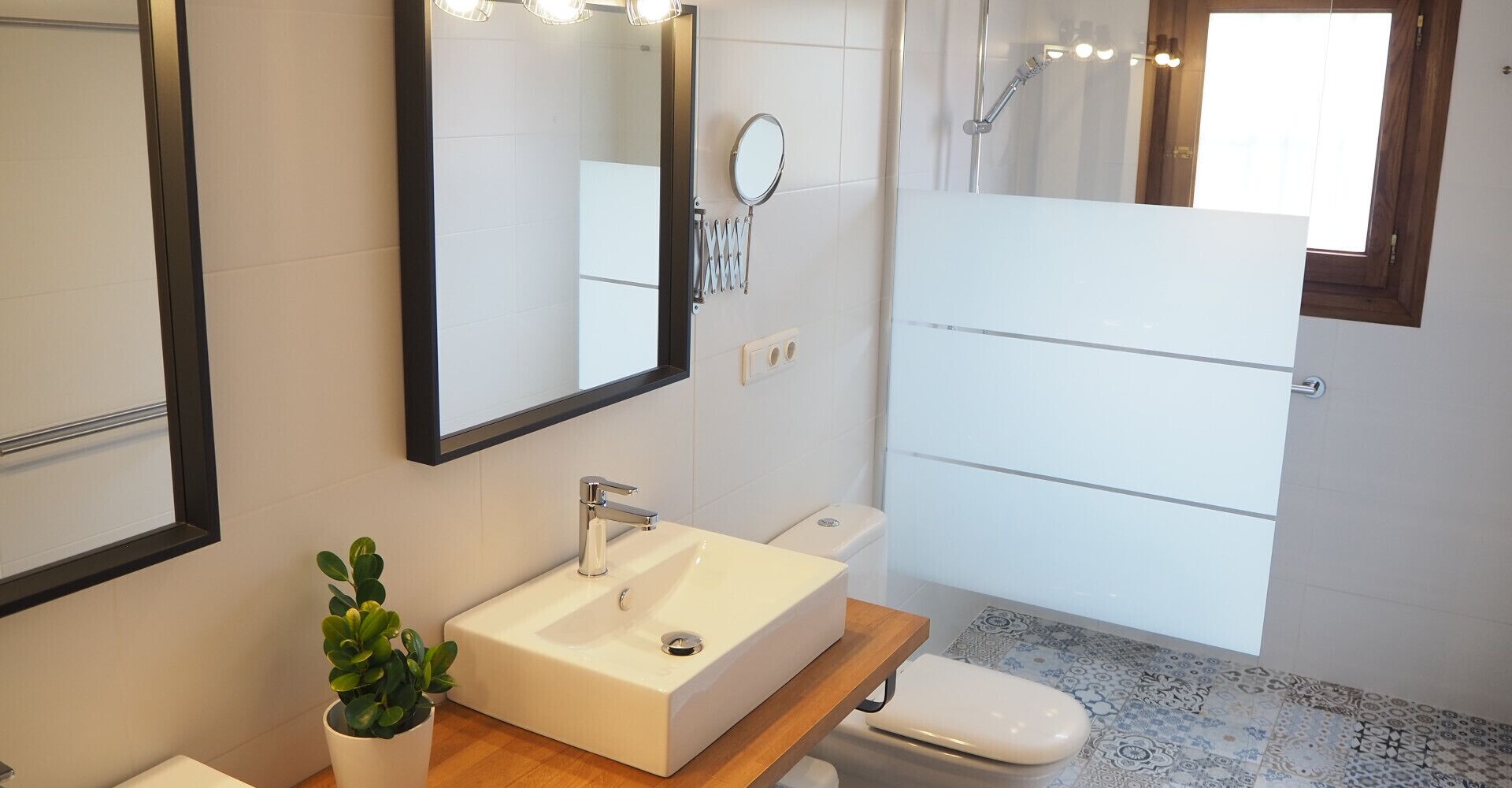 Un baño moderno con un lavamanos, espejo y retrete blancos. Hay una planta pequeña sobre el mostrador del lavamanos y una ducha con puerta de vidrio.