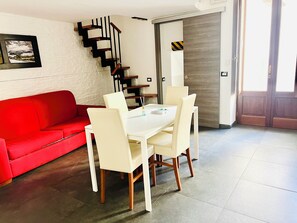 Ein modernes Wohnzimmer mit einem roten Sofa, einem weißen Esstisch mit vier Stühlen, einer Treppe mit schwarzer Handlauf und einer Tür mit Streifenmuster.