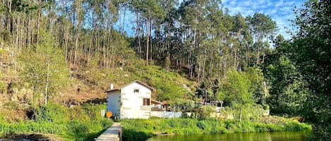 A small white building with a stone foundation, surrounded by lush greenery and a river.