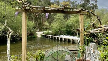 Una pérgola de madera rústica con plantas trepadoras, con vista a un río con un puente y un muro de piedra con una planta en maceta.