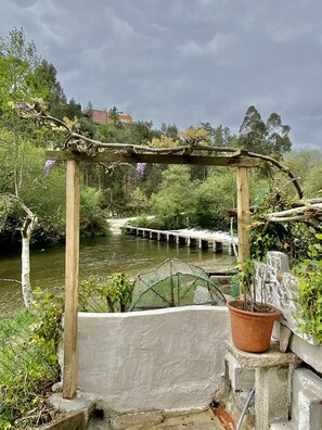 Een rustieke houten pergola met klimplanten, uitkijkend op een rivier met een brug, en een stenen muur met een potplant.