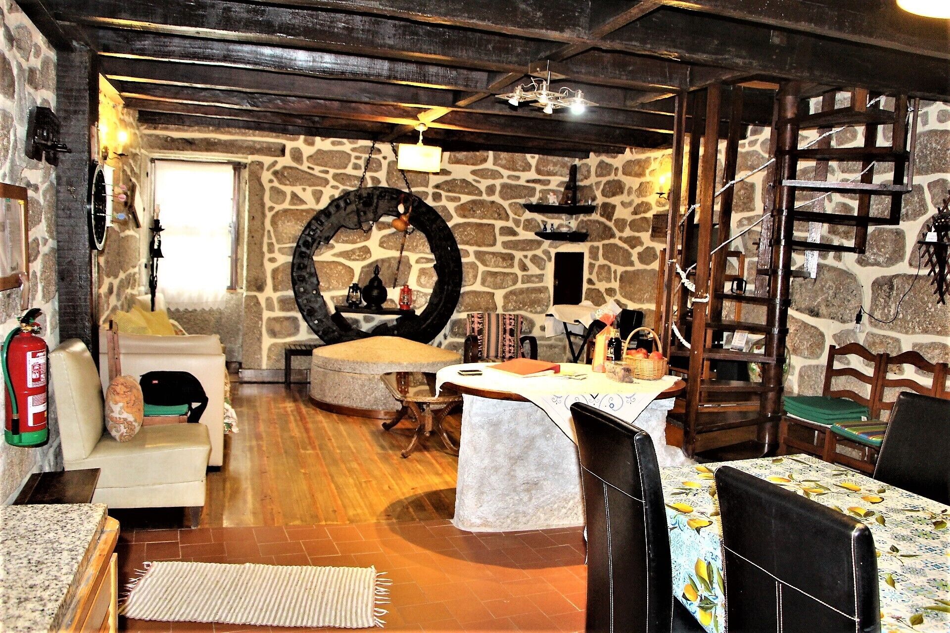 Un interior rústico con paredes de piedra, vigas de madera, una chimenea de piedra, un espejo redondo y un comedor con mesa y sillas.