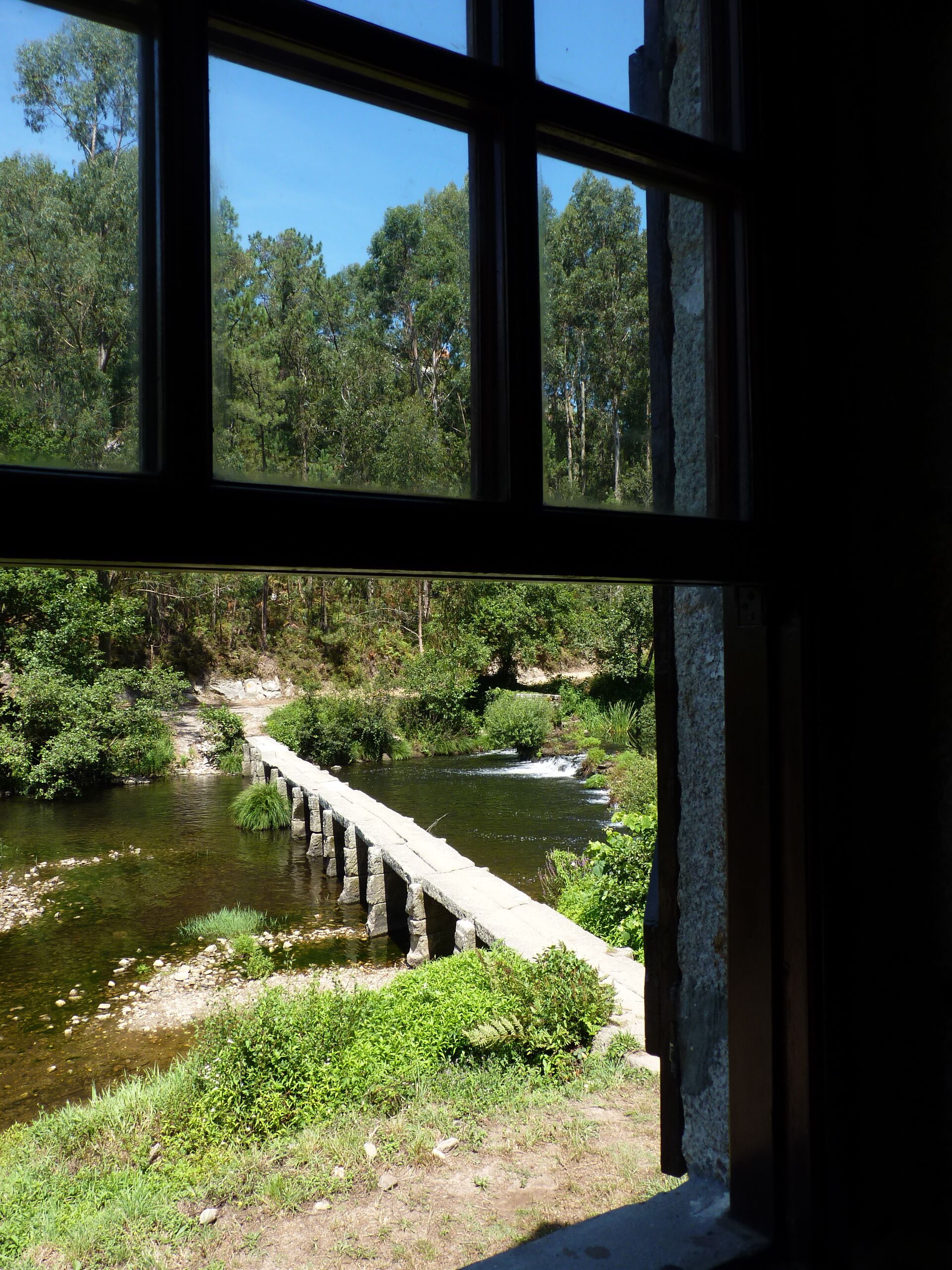 Vista a través de una ventana de un río con un puente de piedra, rodeado de vegetación.