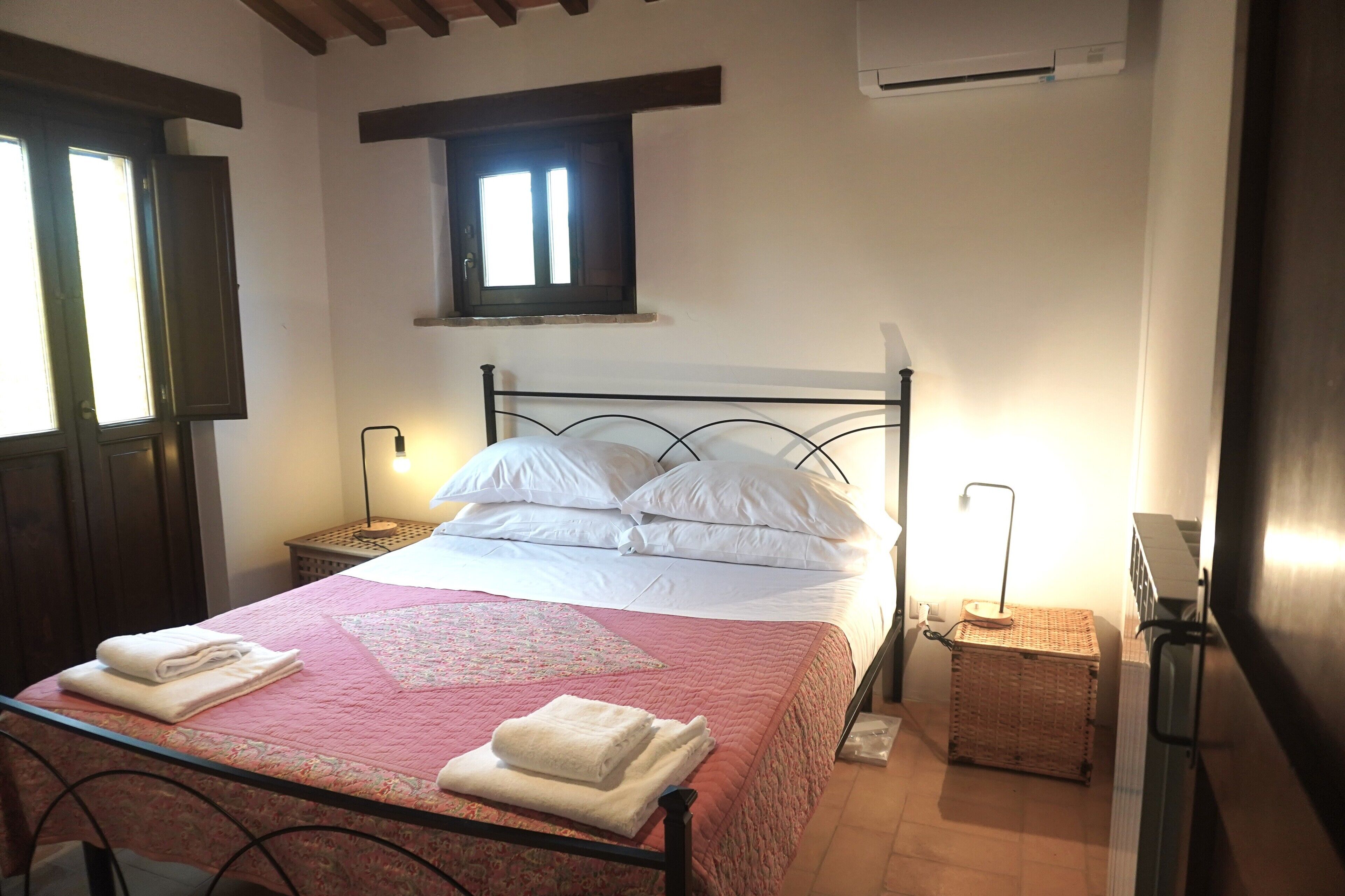 Una cama bien hecha con sábanas blancas y colcha rosa, dos mesitas de noche con lámparas, una ventana con contraventanas y un equipo de aire acondicionado en la pared.
