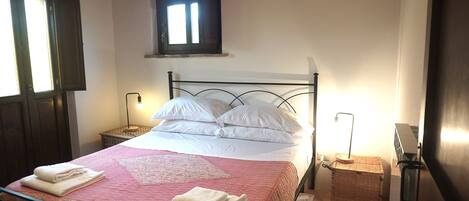 Una cama bien hecha con sábanas blancas y colcha rosa, dos mesitas de noche con lámparas, una ventana con contraventanas y un equipo de aire acondicionado en la pared.