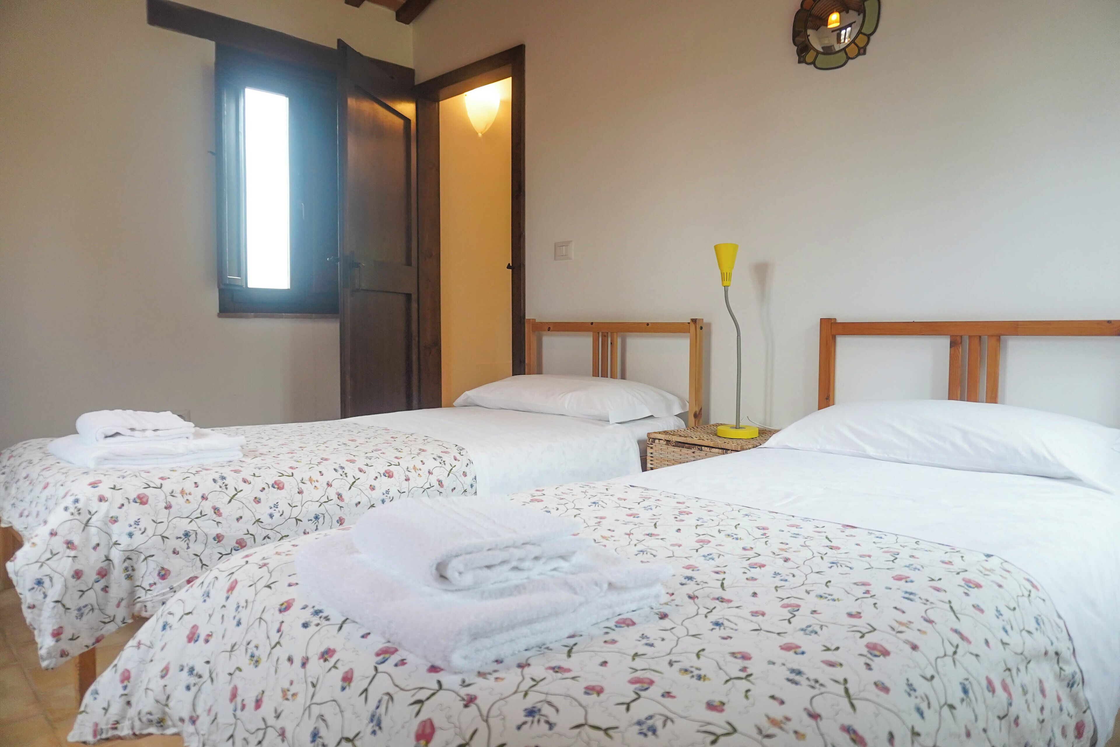 A hotel room with two beds, each with a floral bedspread and white pillows. There is a wooden headboard, a bedside table with a lamp, and a door leading to another room.