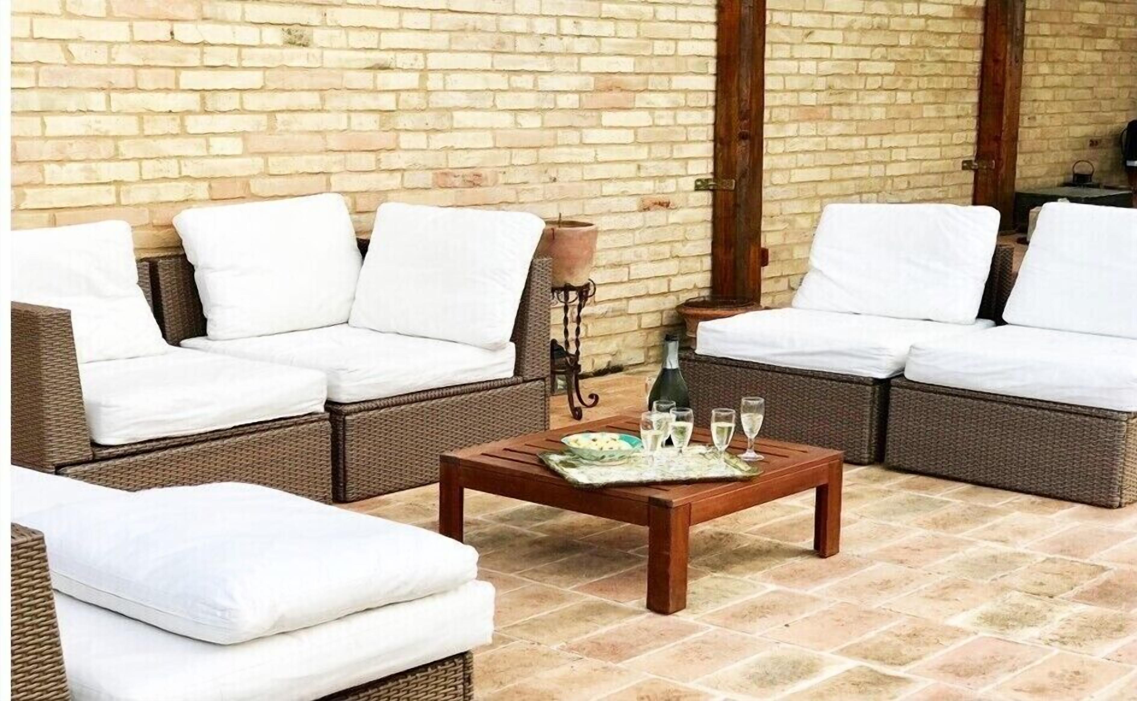 Een knus terras met rieten meubels en witte kussens, een houten bijzettafeltje met glazen en een fles, en een bakstenen muur op de achtergrond.