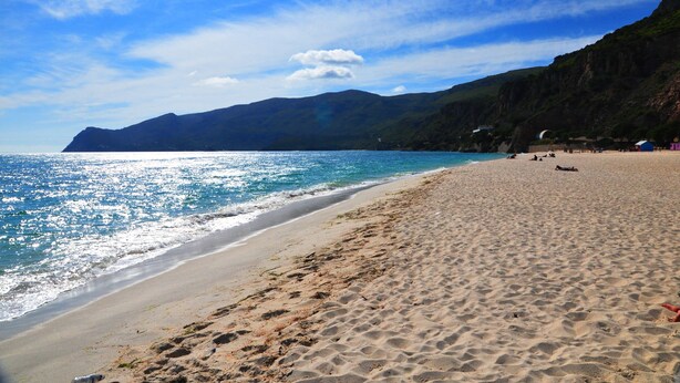 Una playa con aguas azules cristalinas, orilla de arena y montañas al fondo.