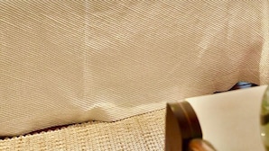 Una cama bien tendida con sábanas blancas y un cojín decorativo. Encima de la cama hay una obra de arte enmarcada y una mesita de noche con una lámpara.