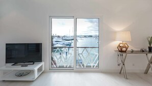 A modern living room with a TV on a white stand, a table with a lamp and a camera, and a view of a marina through a glass door.