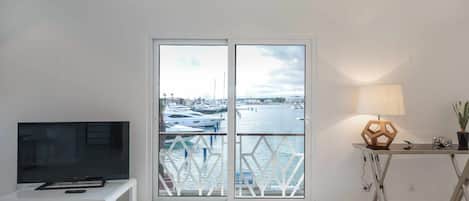 A modern living room with a TV on a white stand, a table with a lamp and a camera, and a view of a marina through a glass door.