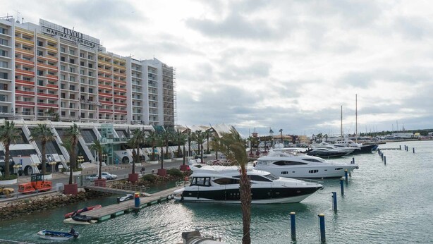 A marina with several yachts docked, a modern building with balconies, and palm trees along the waterfront.