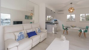 A modern living room with a sofa, a glass dining table, and chairs. There is a kitchen area in the background.