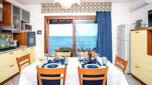 Un coin repas avec une table dressée pour deux personnes, une vue sur la mer par une porte vitrée et une cuisine équipée d’un four à micro-ondes et d’un réfrigérateur.