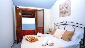 Une chambre avec un lit, une table de chevet, une lampe, une œuvre d’art représentant une étoile de mer, une fenêtre donnant sur la mer et un plateau de petit-déjeuner.