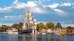 Een historisch zeilschip aangemeerd aan een haven, met traditionele gebouwen en herfstkleuren op de achtergrond.