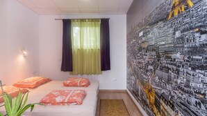 Une chambre à coucher avec un lit, des oreillers, une plante en pot, une fenêtre avec des rideaux et une fresque représentant un paysage urbain sur le mur.