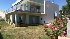 Une maison moderne à deux étages, dotée d’un balcon, d’un porche abrité et d’un jardin où poussent des rosiers roses.