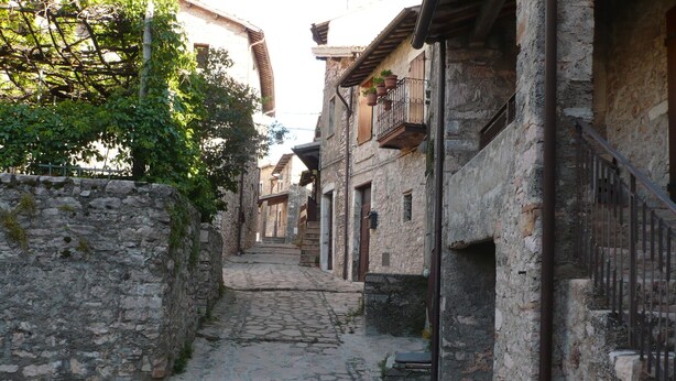 Una estrecha calle adoquinada bordeada por edificios de piedra con balcones, una pared de piedra a un lado y una cerca metálica al otro.