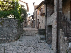 A narrow cobblestone street flanked by stone buildings with balconies, a stone wall on one side, and a metal fence on the other.