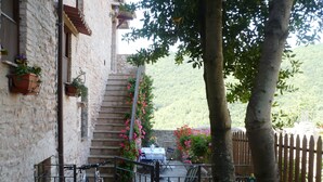 Un edificio de piedra con una escalera que lleva a una terraza. Hay una bicicleta aparcada cerca de un portón, y una mesa con sillas en la terraza.
