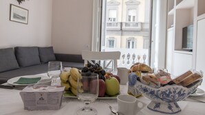 Una mesa de comedor con frutas, pan y un bol de pan, una silla blanca, y una ventana con vistas a edificios.