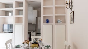 Un comedor con una mesa preparada para cuatro personas, una pared blanca con un jarrón azul y una cocina al fondo.