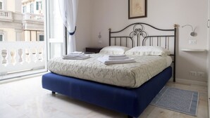 Un dormitorio con cama, mesita de noche, ventana con vistas a edificios y un cuadro colgado en la pared.