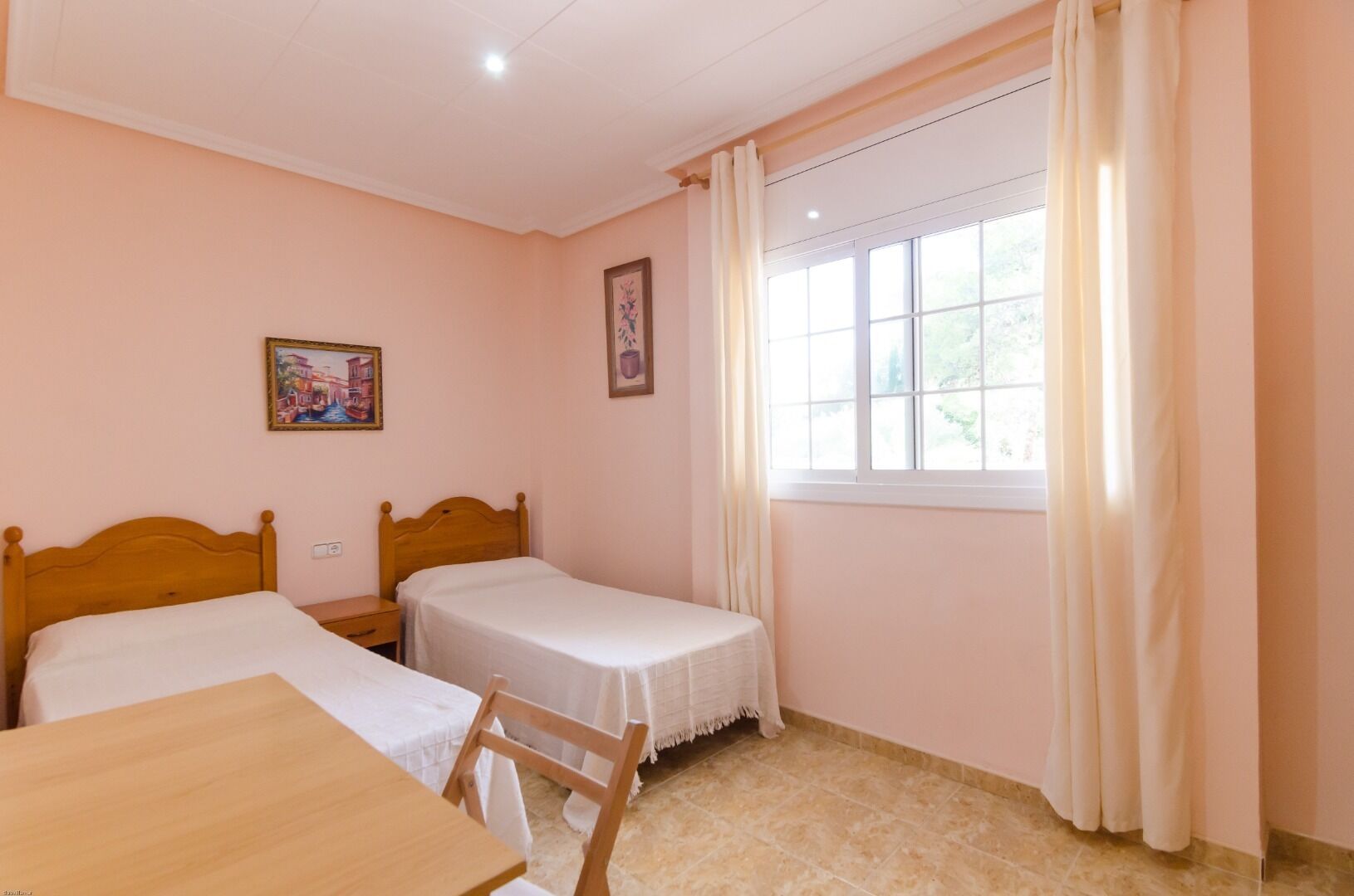 A room with two single beds, a wooden table, and a window with curtains.