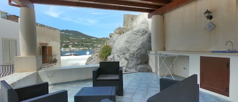 Un patio al aire libre con mobiliario de mimbre, piso de piedra, vista al mar y un edificio con fachada blanca.