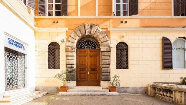 Un edificio con una porta centrale in legno, due piante in vaso su entrambi i lati, e una insegna con scritto 'Coldwell Banker' a sinistra.