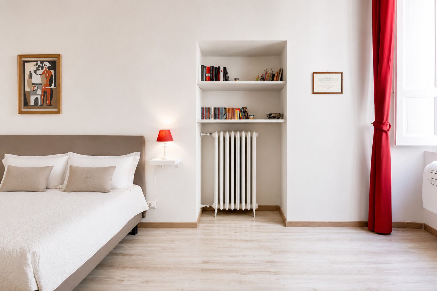 A bedroom with a bed, a bedside table with a lamp, a radiator, a bookshelf, and a window with curtains.