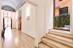 Un passadís ampli amb terres de fusta, una escala que condueix a un balcó amb vistes a un bosc frondós, i una zona de menjador amb cadires i una taula.