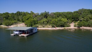 Une maison flottante dotée d’une terrasse couverte et d’un espace salon, entourée d’eau et d’une forêt dense.